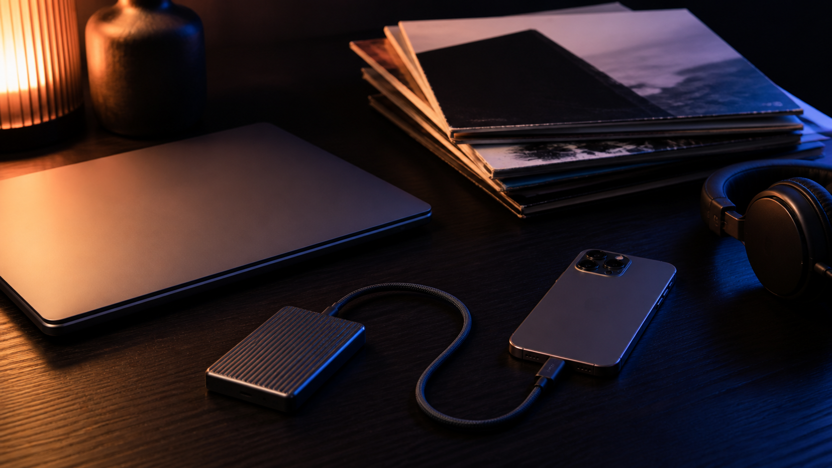A laptop, portable drive and iPhone connected by cable on a dark desk beside a stack of record sleeves.