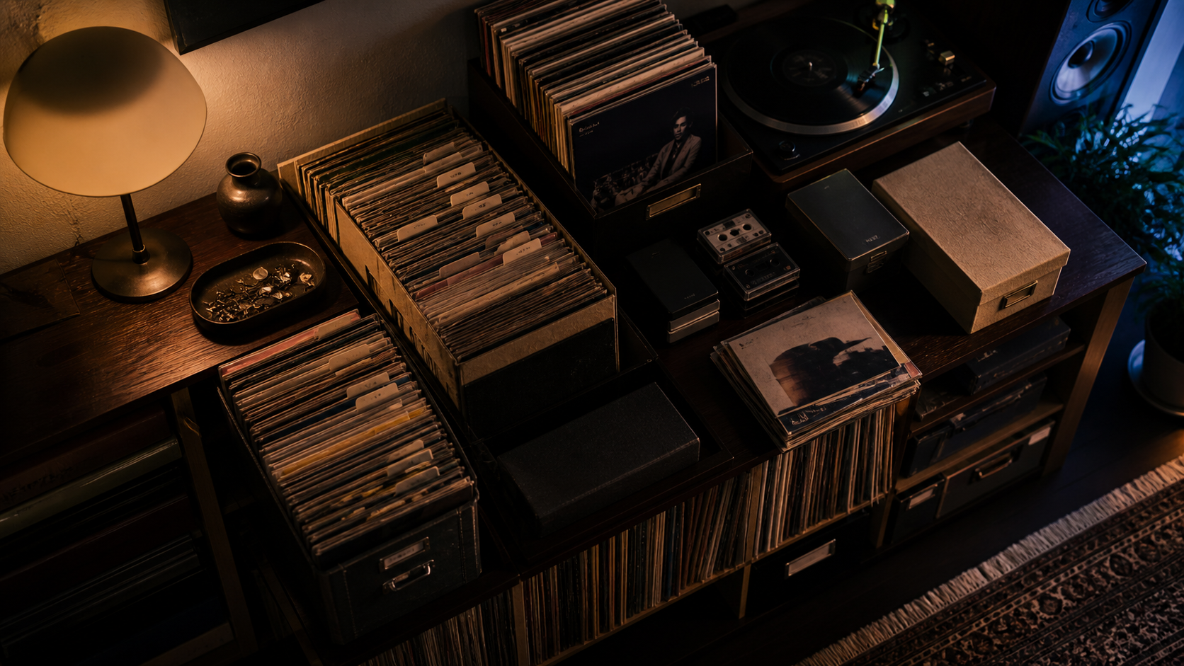 A warmly lit sideboard holding filed records, CDs, tapes and a turntable in a home listening room.